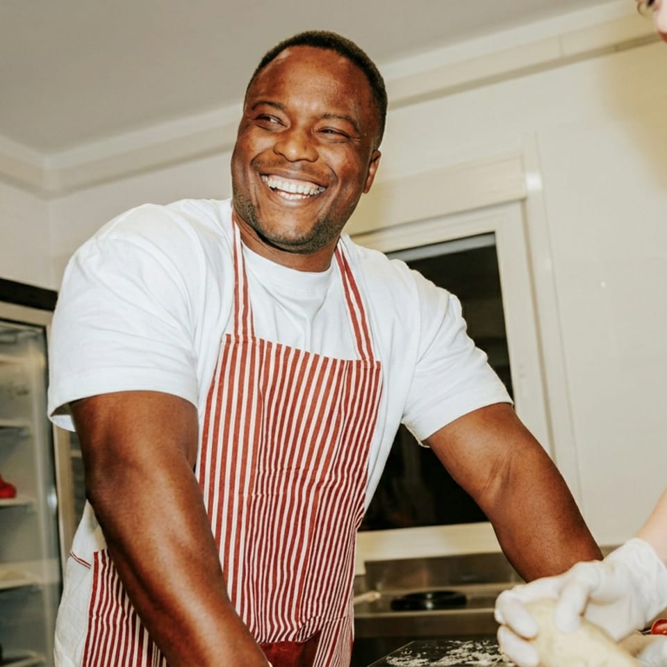 A smiling chef in a red and white striped apron works with another person in a kitchen.