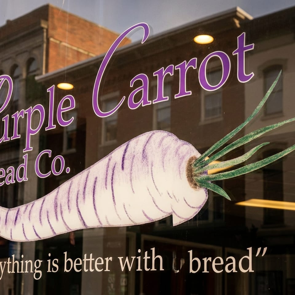 Purple Carrot Bread Co. logo and slogan on a glass window reflecting a brick building.